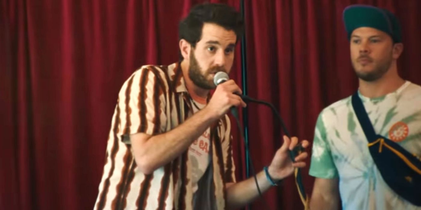 Theater Camp: Angelo is on stage with the mic with a Tory in a tie dye shirt behind him