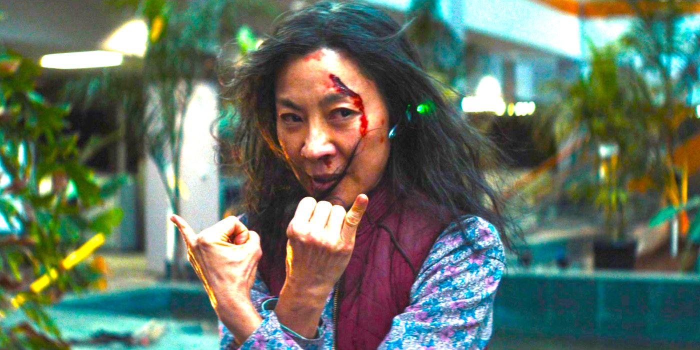 Michelle Yeoh in Everything Everywhere All At Once in a mall making hand gestures with here pinky fingers sticking up, her hair mussed and her face gashed open
