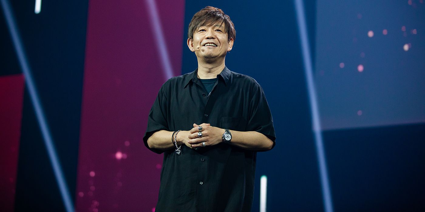 Yoshi-P stands on stage in front of a blue background. He wears a black button-down with a black t-shirt underneath, and a headset microphone.