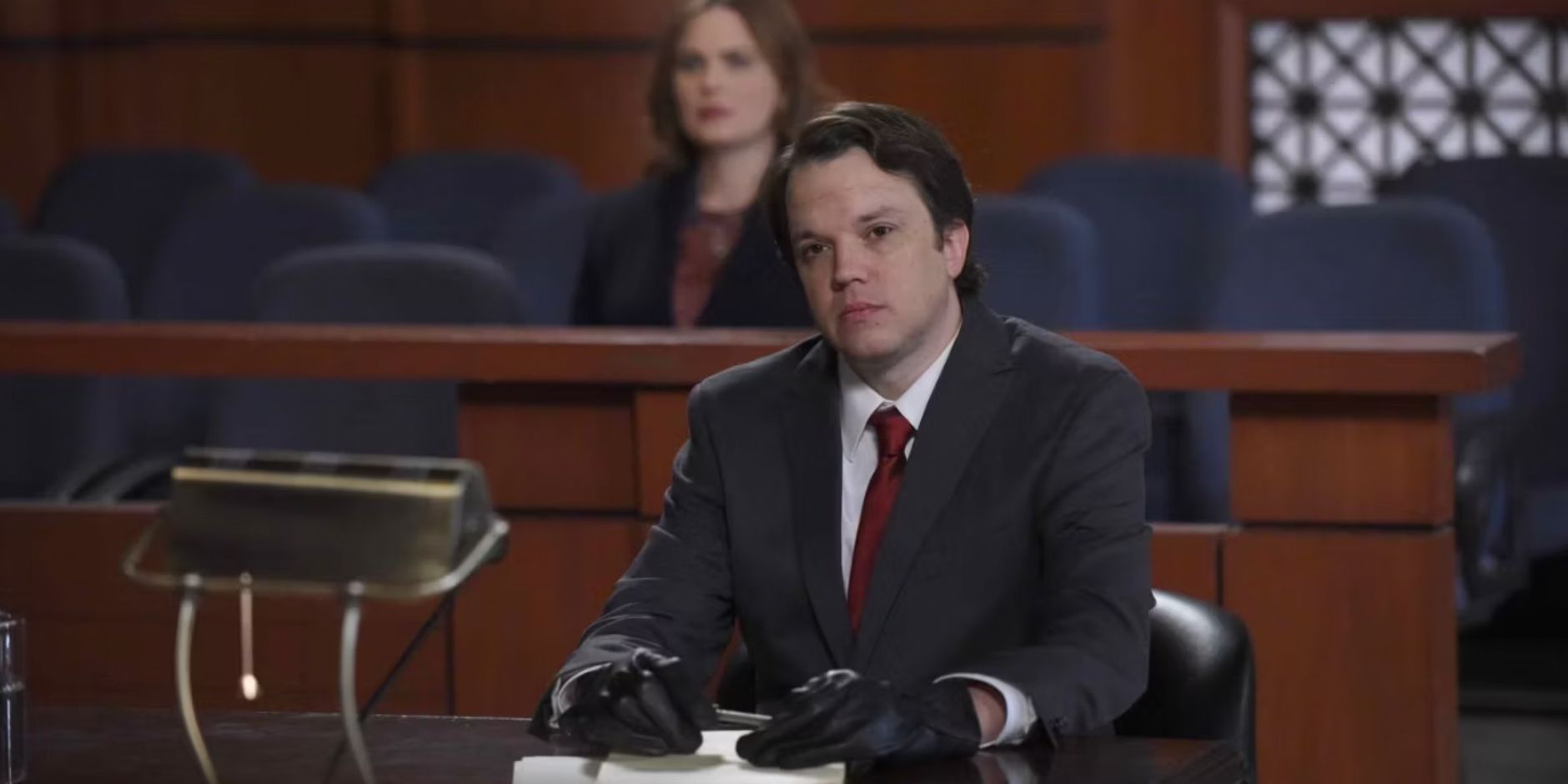 Zack sitting in a courtroom with Brennan behind him in Bones