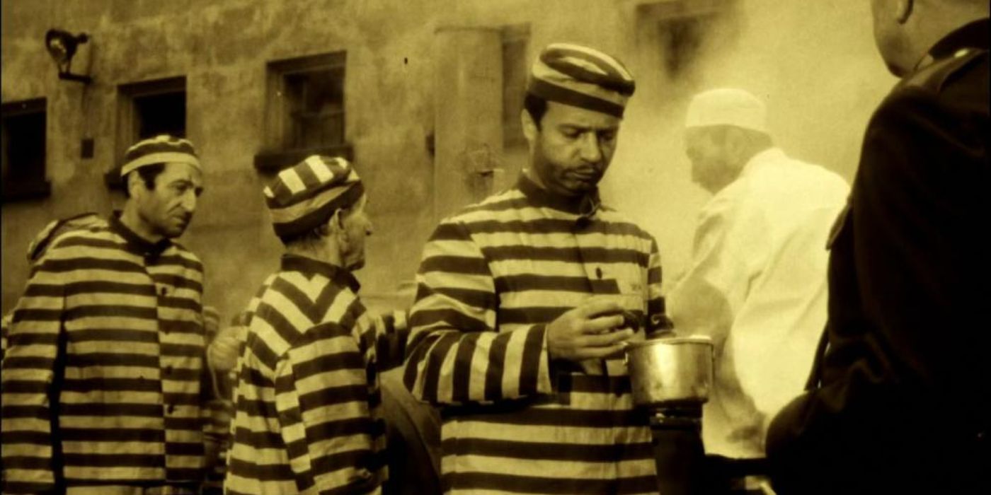 A prisoner broodingly looking at his vessel while standing in line with other prisoners in pin-striped uniforms in Happy End