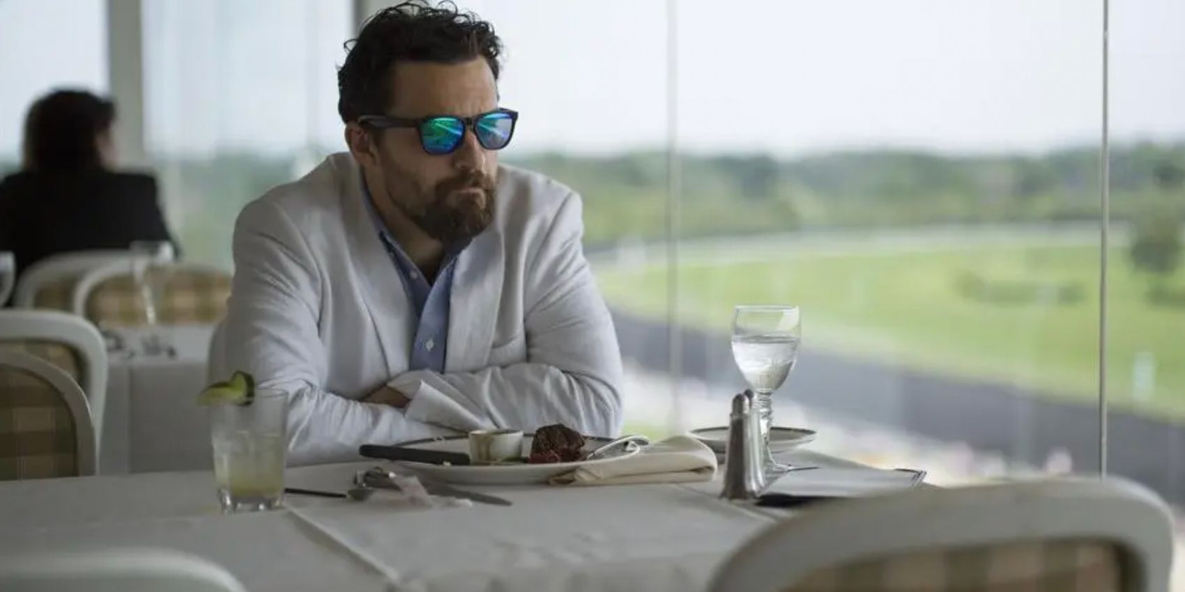 Jake Johnson in white and wearing sunglasses at a table in Win It All