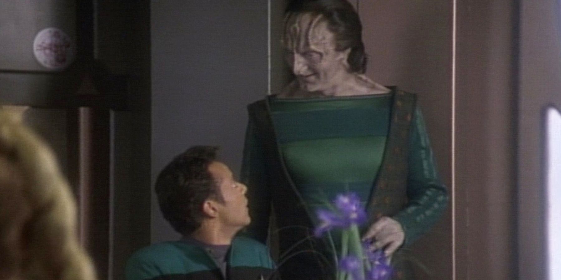 A young Dr. Bashir (portrayed by Alexander Siddig,) looks up at Garak (portrayed by Andrew Robinson,) in a clearly off-kilter way as Garak looks smug. Siddig has tan skin and short brown hair, and wears a blue and black uniform. Garak has grey skin, ridges on his forehead and around his eyes that show he's a Cardassian, and nearly shoulder-length black hair. Garak wears a blue-and-green striped top with a brown vest.