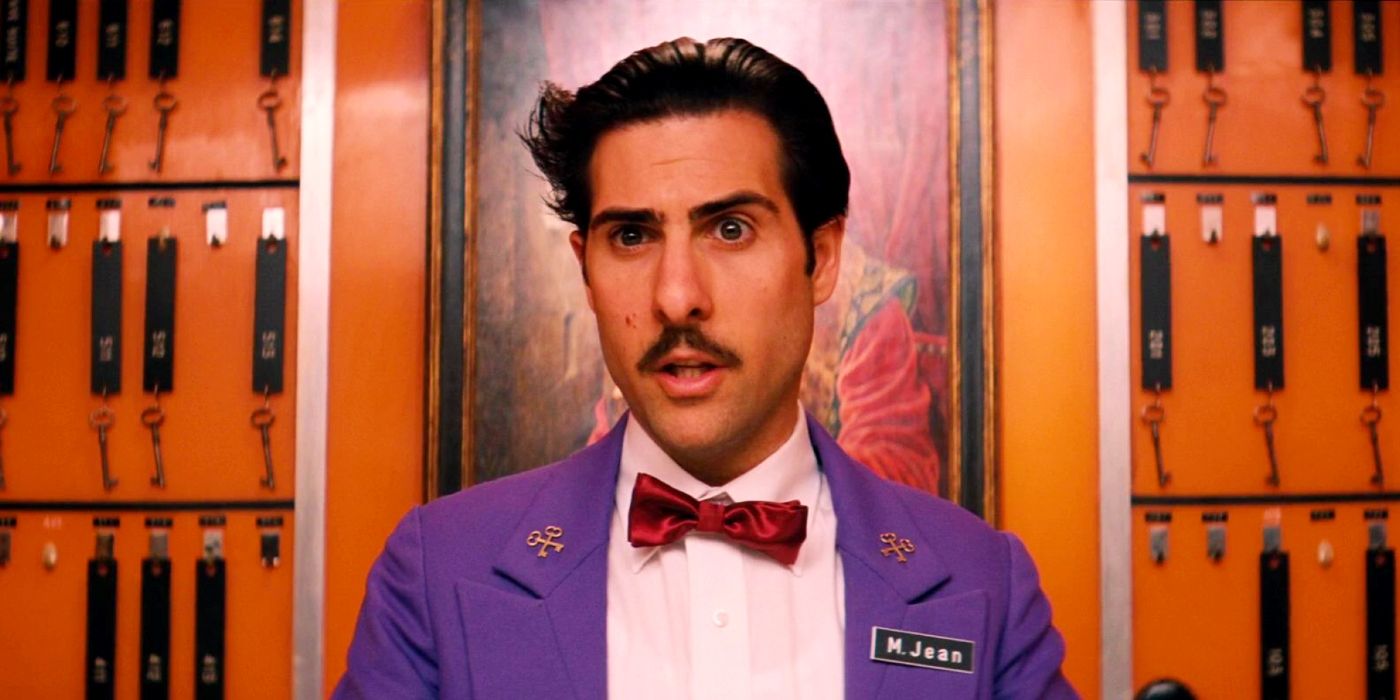 Jason Schwartzman standing in front of a wall of room keys in The Grand Budapest Hotel.