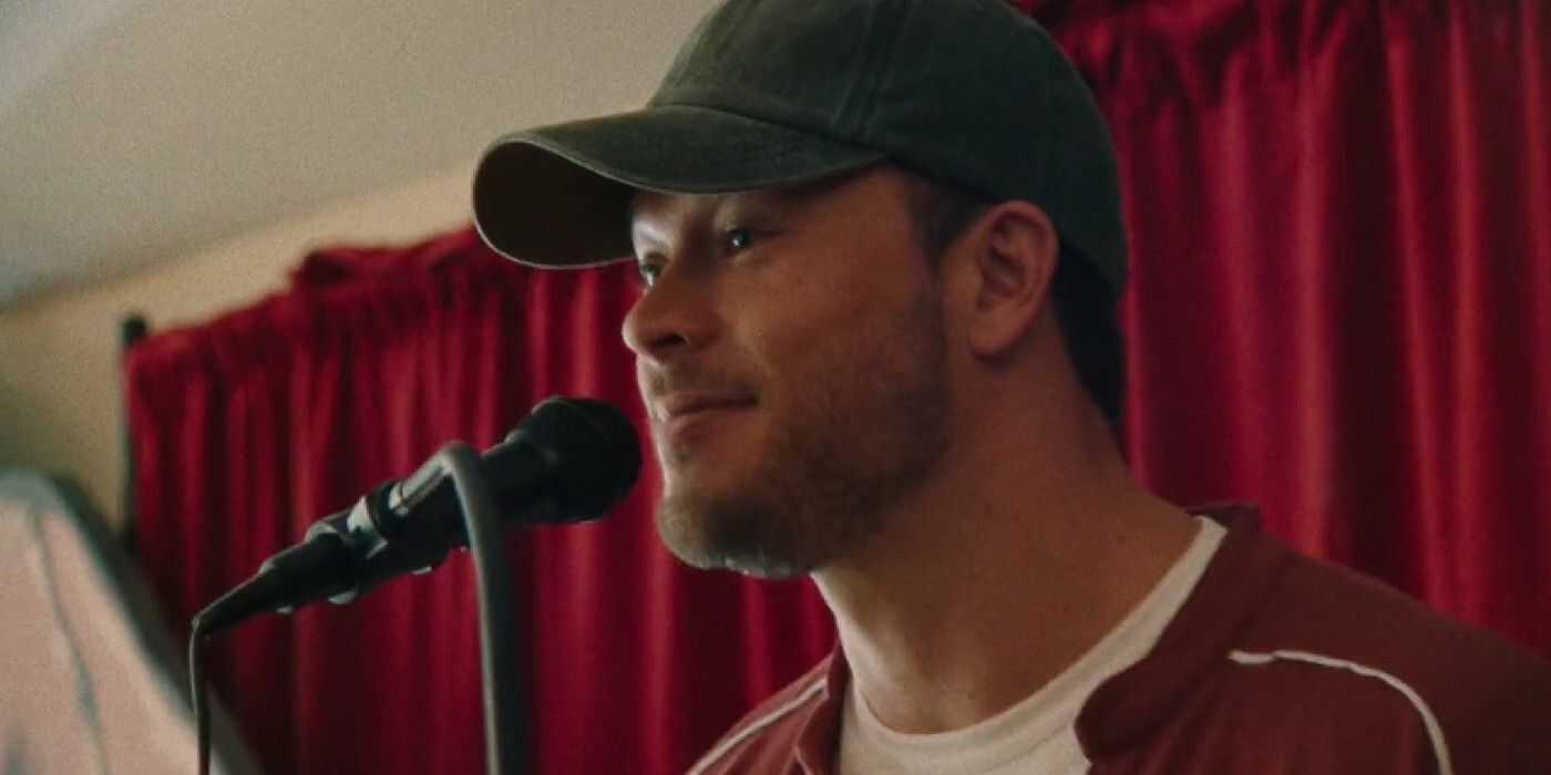 Jimmy Tatro as Troy in Theater Camp, wearing a dark-colored hat and maroon jacket with a white shirt as he smirks in front of a microphone.