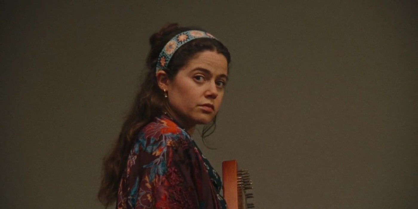 A screencap of Molly Gordon as Rebecca-Diane in Theater Camp. She's looking to the side and holding an instrument.