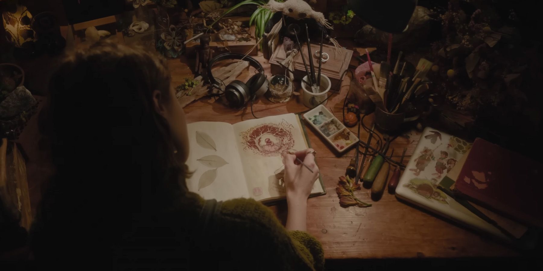 Woman drawing in a green book open on a cluttered desk.