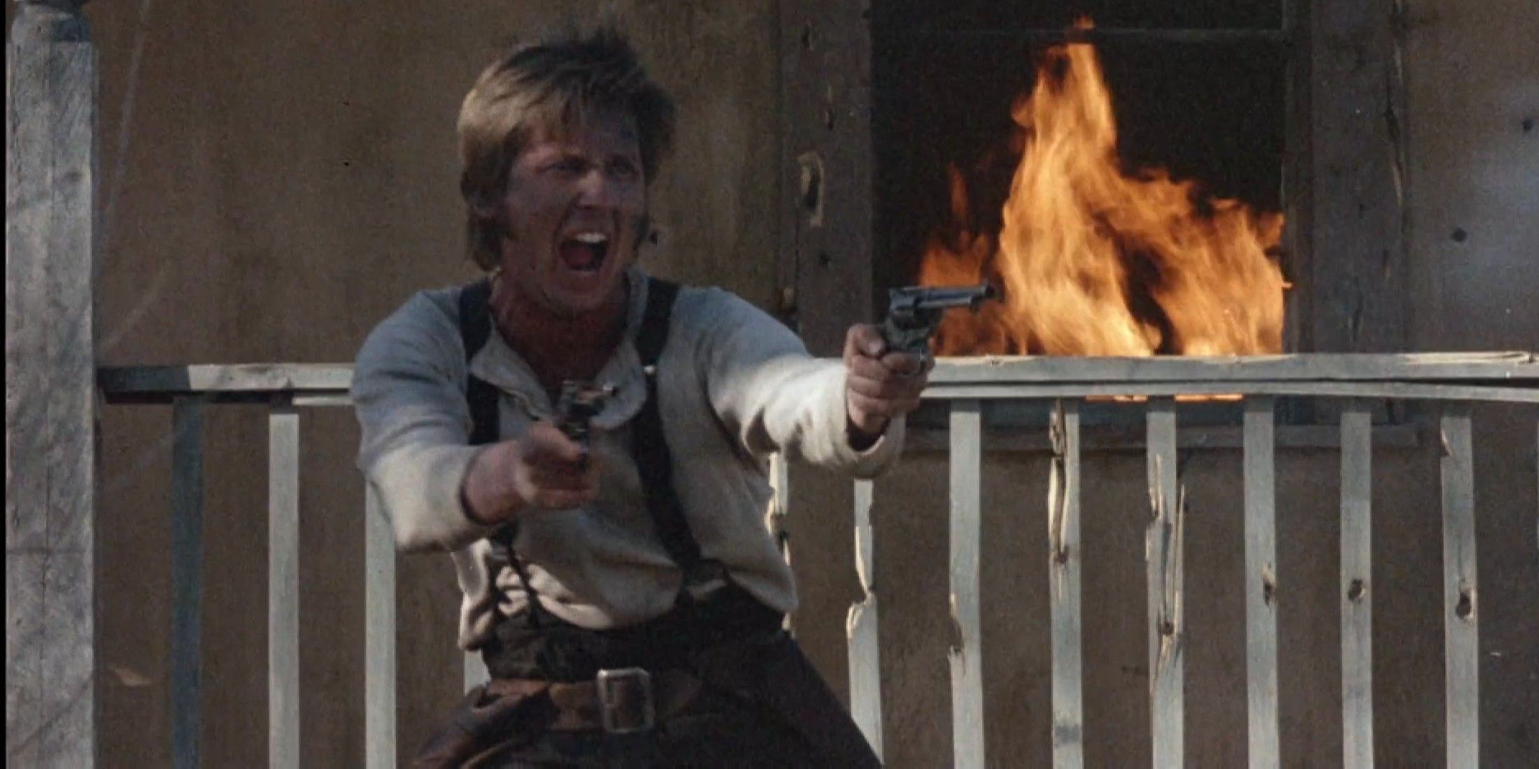 Emilio Estevez como Billy the Kid disparando duas armas do lado de fora de uma casa em chamas em Young Guns