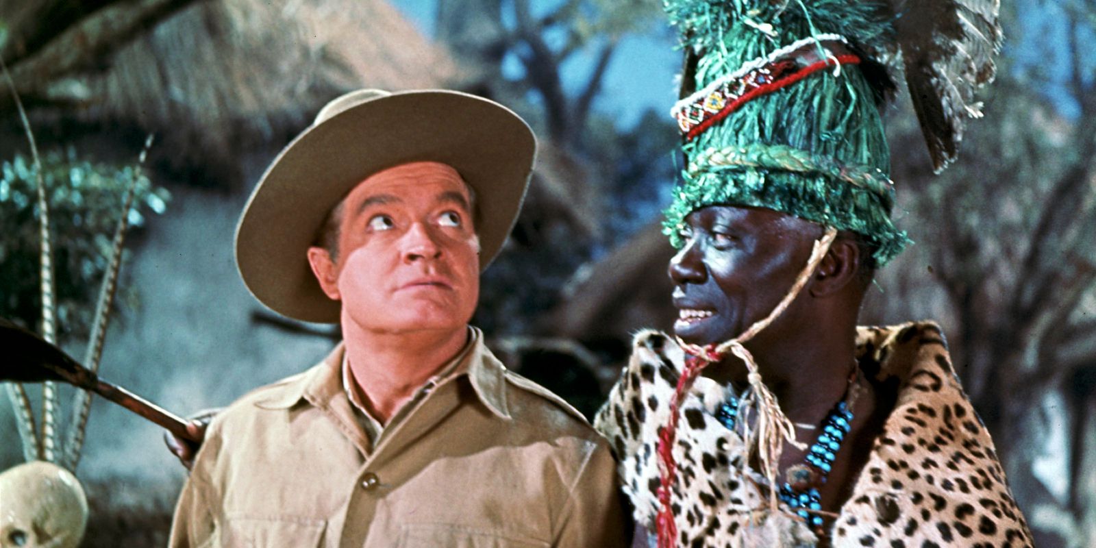 Two men stand together in Africa with a hut in the background.