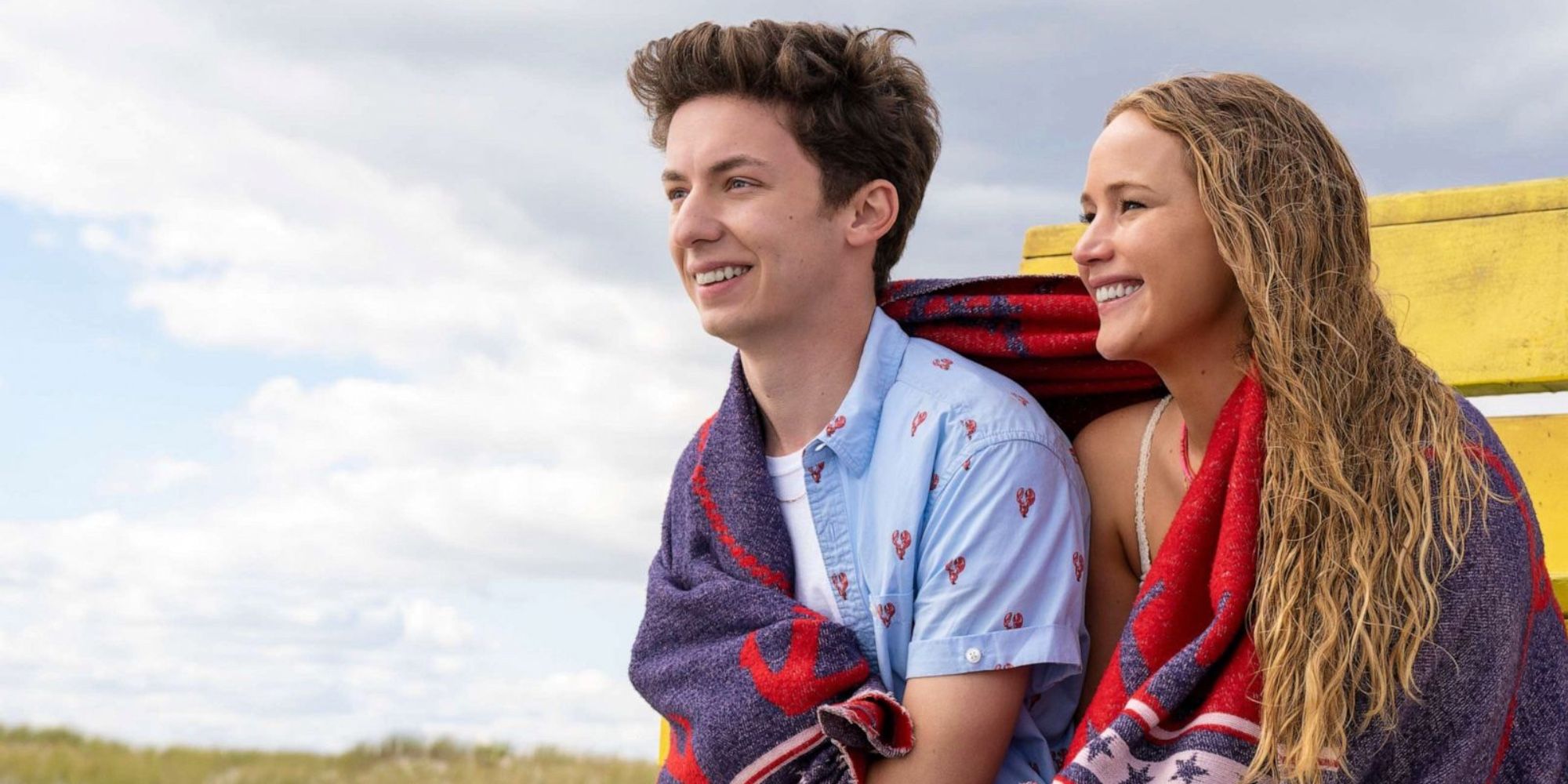 Maddie (Jennifer Lawrence) and Percy (Andrew Barth Feldman) have a towel around them, while they sit at the beach in No Hard Feelings.