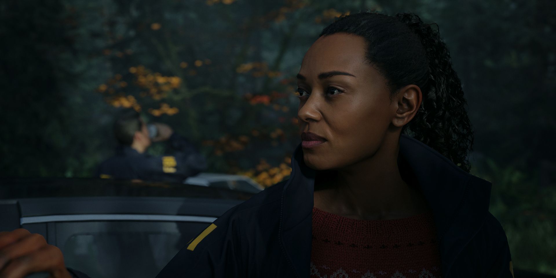 Saga, standing in front of a squad car, looks to her right with a vague smile in a screenshot from Alan Wake 2.