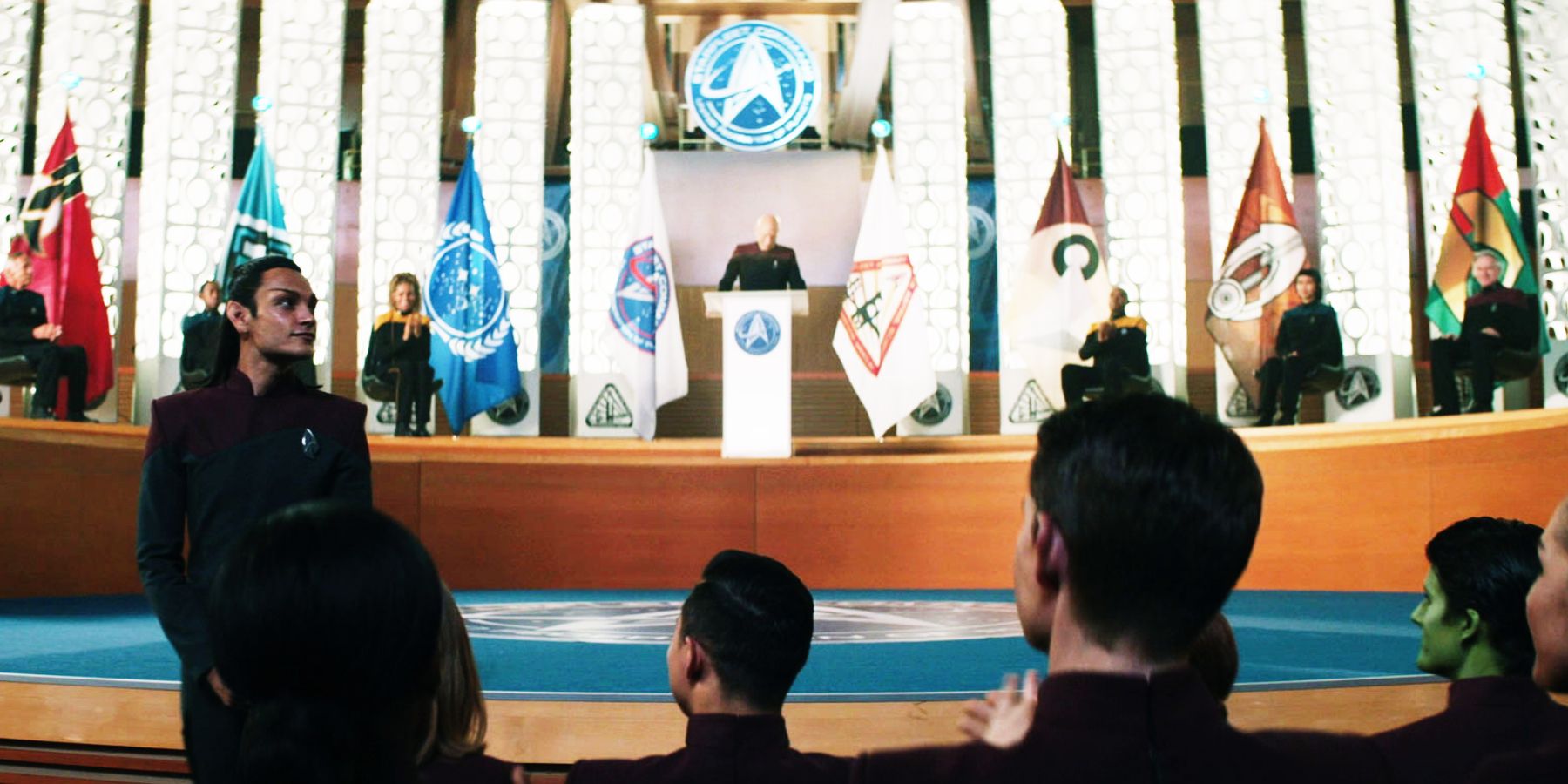 A student receives applause at a graduation ceremony, with various flags on display behind them