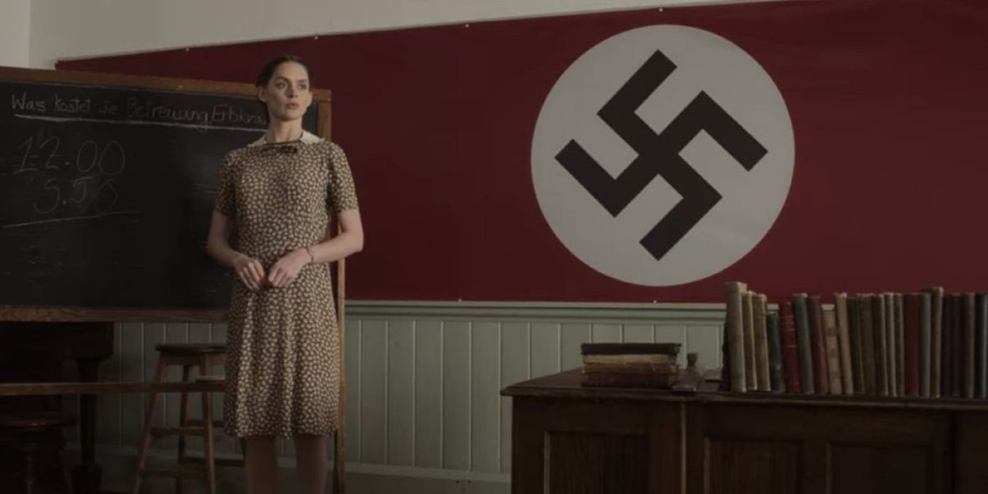 A female teacher standing in front of a classroom with a Nazi symbol on the wall in Forgive Us Our Trespasses