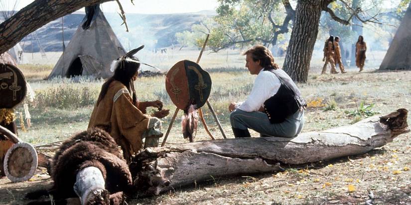 Dunbar sitting on a log talking to a Native American character in Dances With Wolves