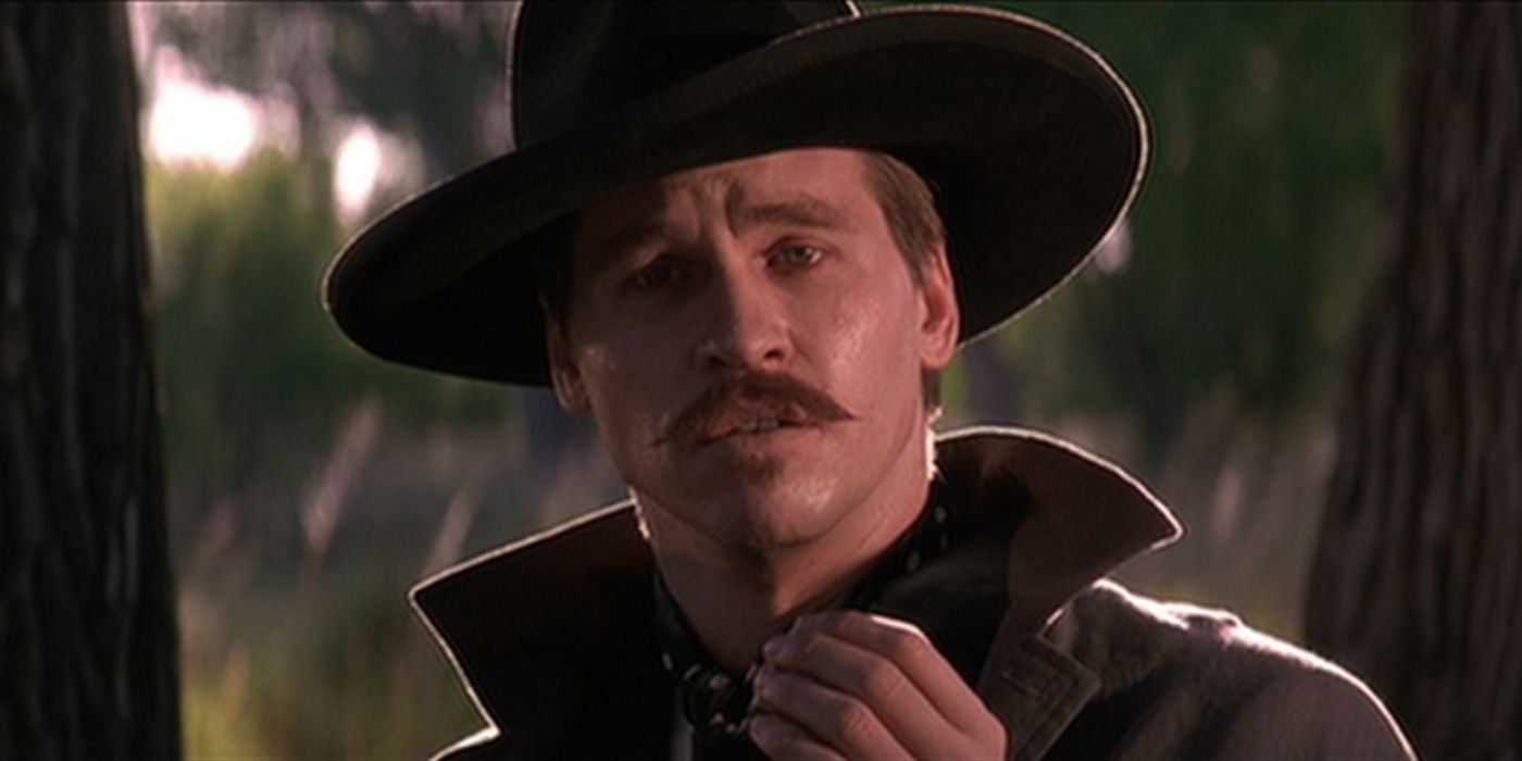 Val Kilmer as Doc Holliday holding his hand up and speaking with the Earp brothers in 1993's Tombstone​​​​​​​.