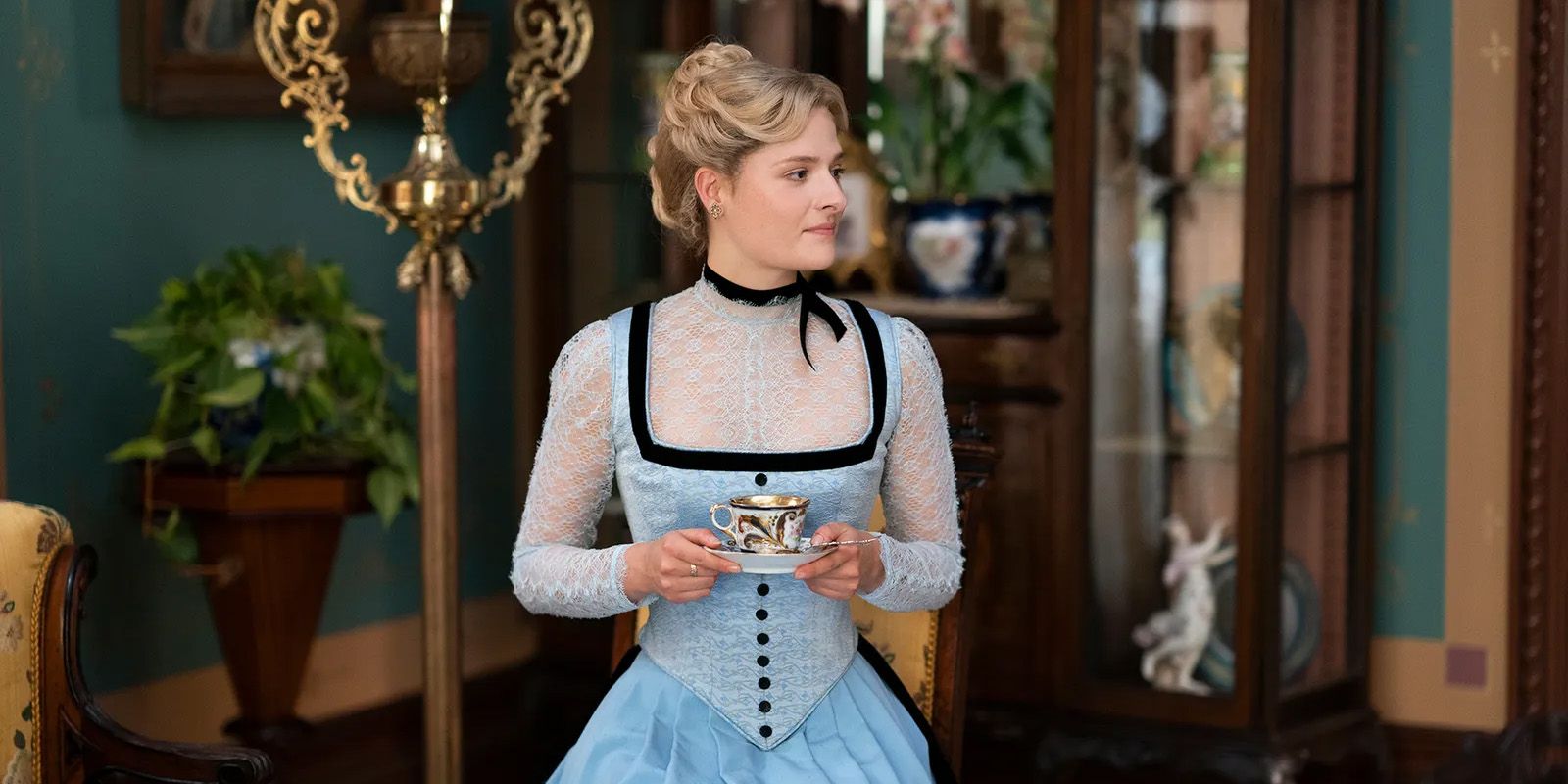 Louisa Jacobson as Marian in the Gilded Age. She's holding a cup of tea and wearing a blue and black dress with white lace on the top