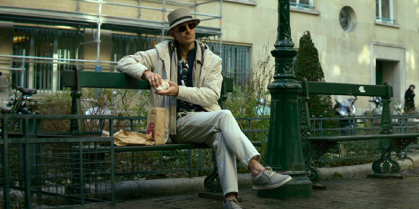 Michael fassbender eats a McMuffin sitting on a bench in the killer