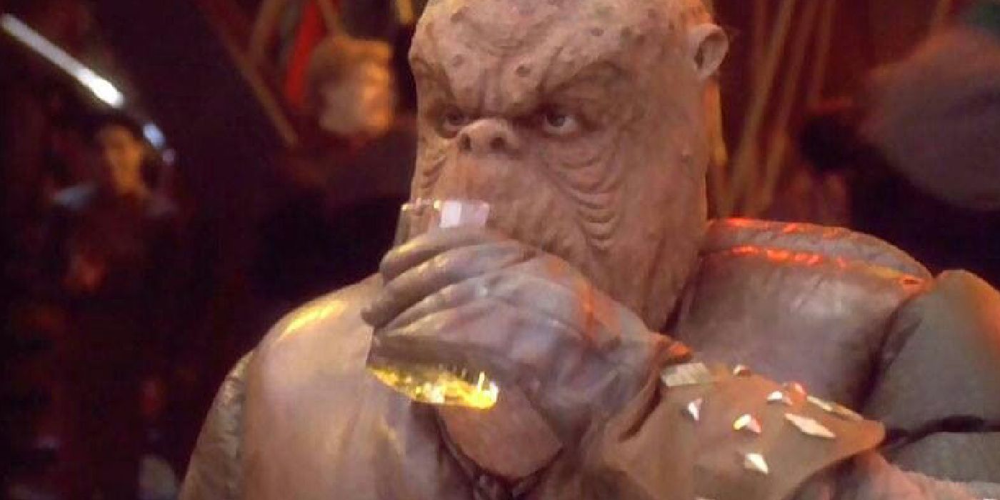A large alien drinks what looks like whisky out of a square glass