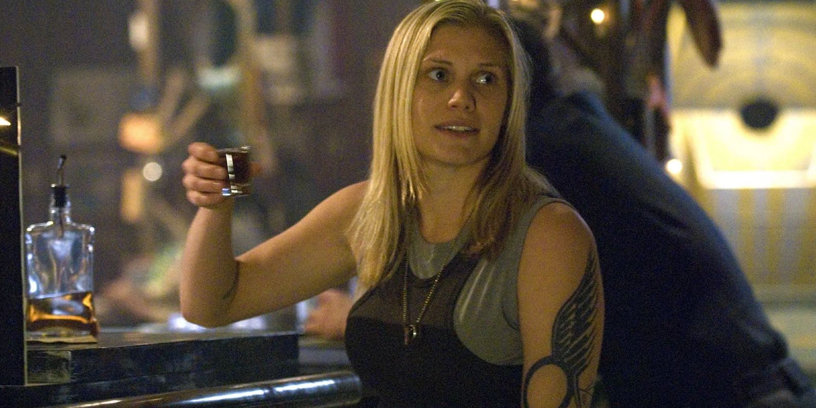 Starbuck (Katee Sackhoff) levanta um copo no ar enquanto está sentado em um bar na Battlestar Galactica