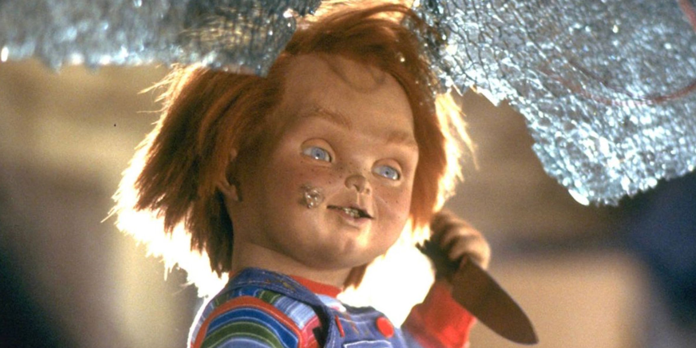 Chucky holding a knife in front of broken glass in Childs Play 2