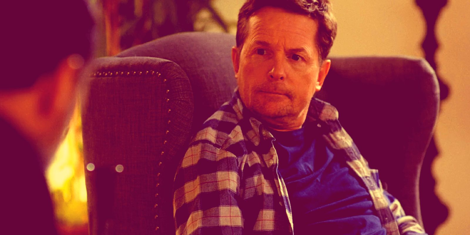 Michael J. Fox sits in a chair looking over at another man in Curb Your Enthusiasm.