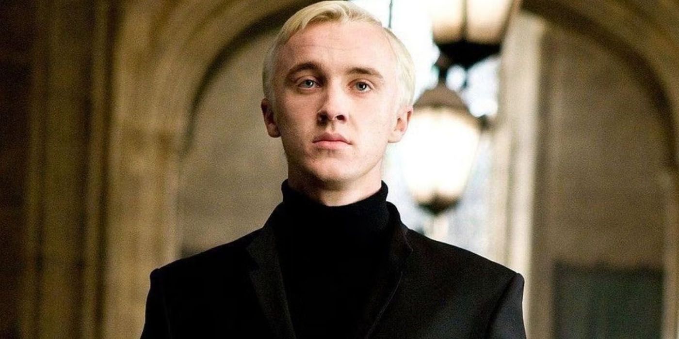 Tom Felton as Draco Malfoy wearing all black and standing in Hogwarts hall in Harry Potter