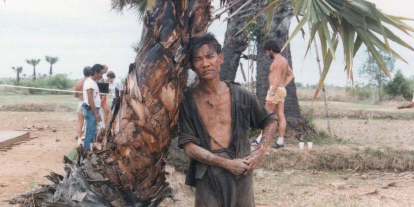 Haing S. Ngor in costume as Dith Pran leans against a palm tree on set of The Killing Fields.