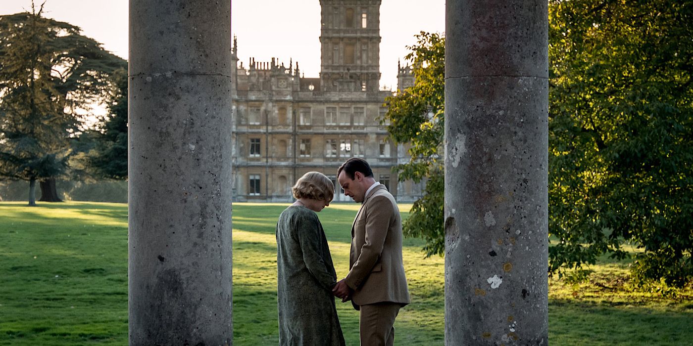 Edith and Bertie stand outside between two pillars in front of a large estate in Downton Abbey.