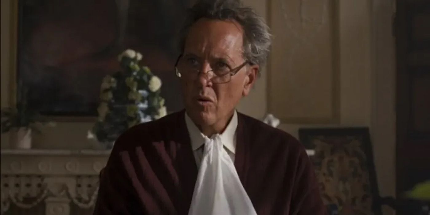 Sir James Catton (Richard E. Grant) sitting at the table, wearing glasses, with a napkin tucked in his shirt in Saltburn.