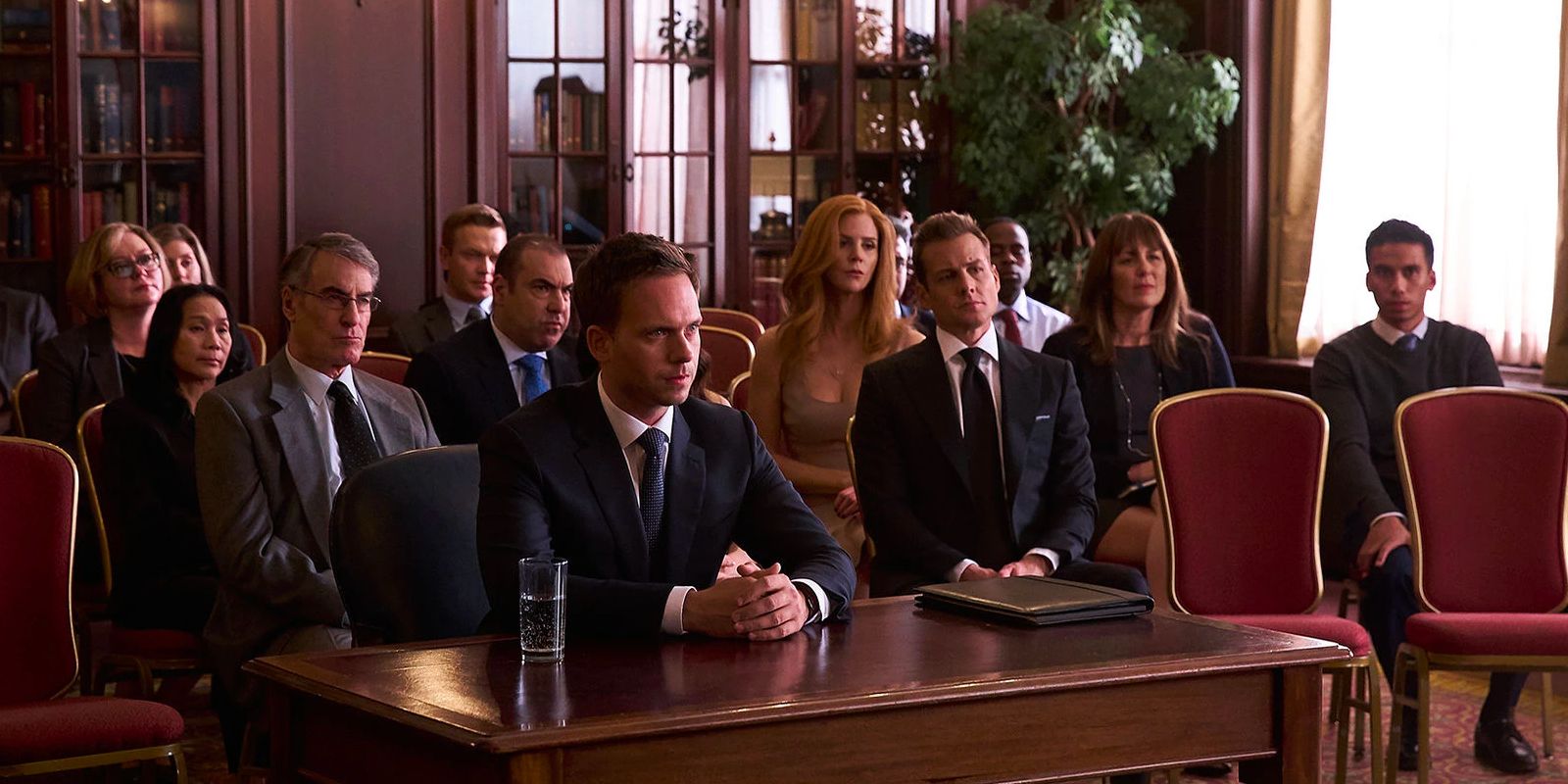 Mike Ross sitting at a desk surrounded by the cast of Suits during character and fitness.