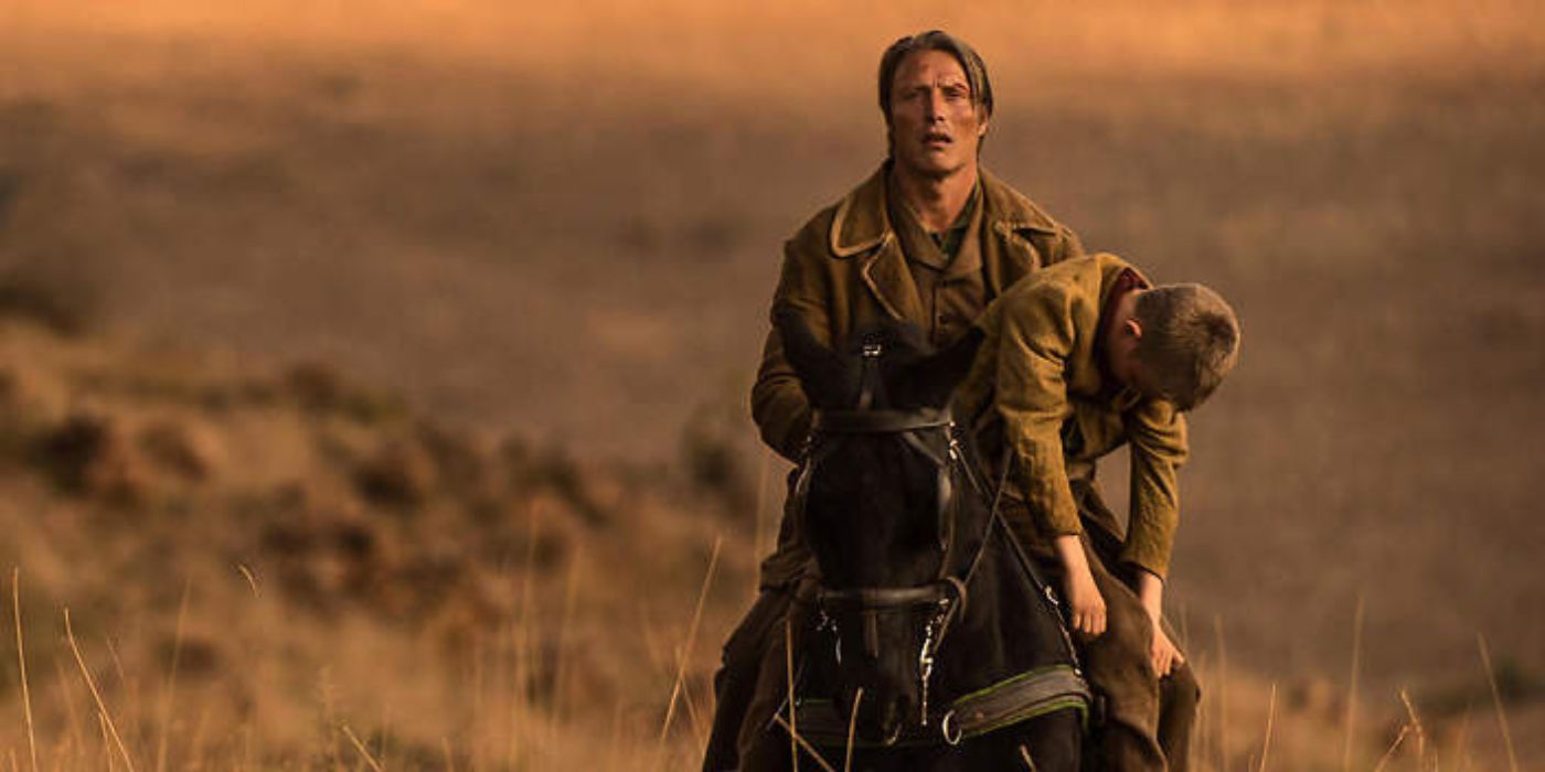 Jon Jensen (Mads Mikkelsen) wearily rides on horseback with a kid slumped over his arm in The Salvation