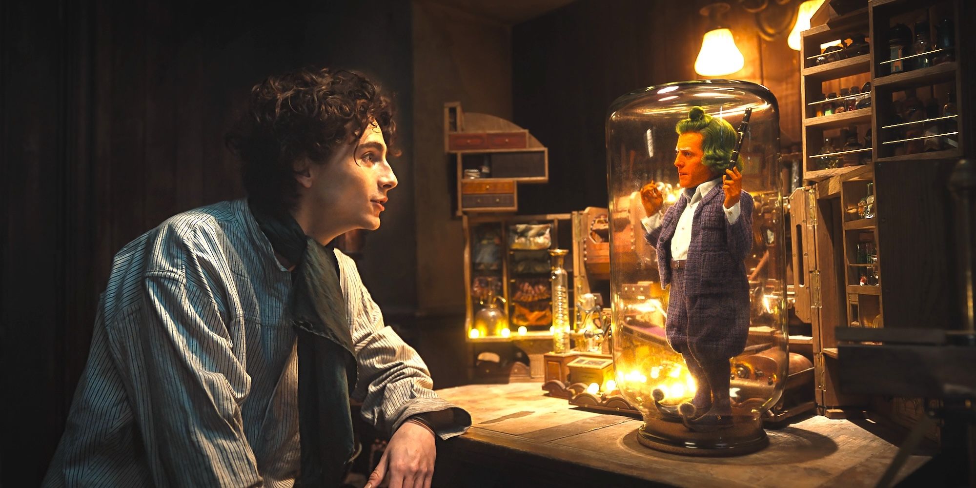 Willy Wonka (Timothée Chalamet) looking curiously at an Oompa Loompa (Hugh Grant) in a glass enclosure in Wonka.
