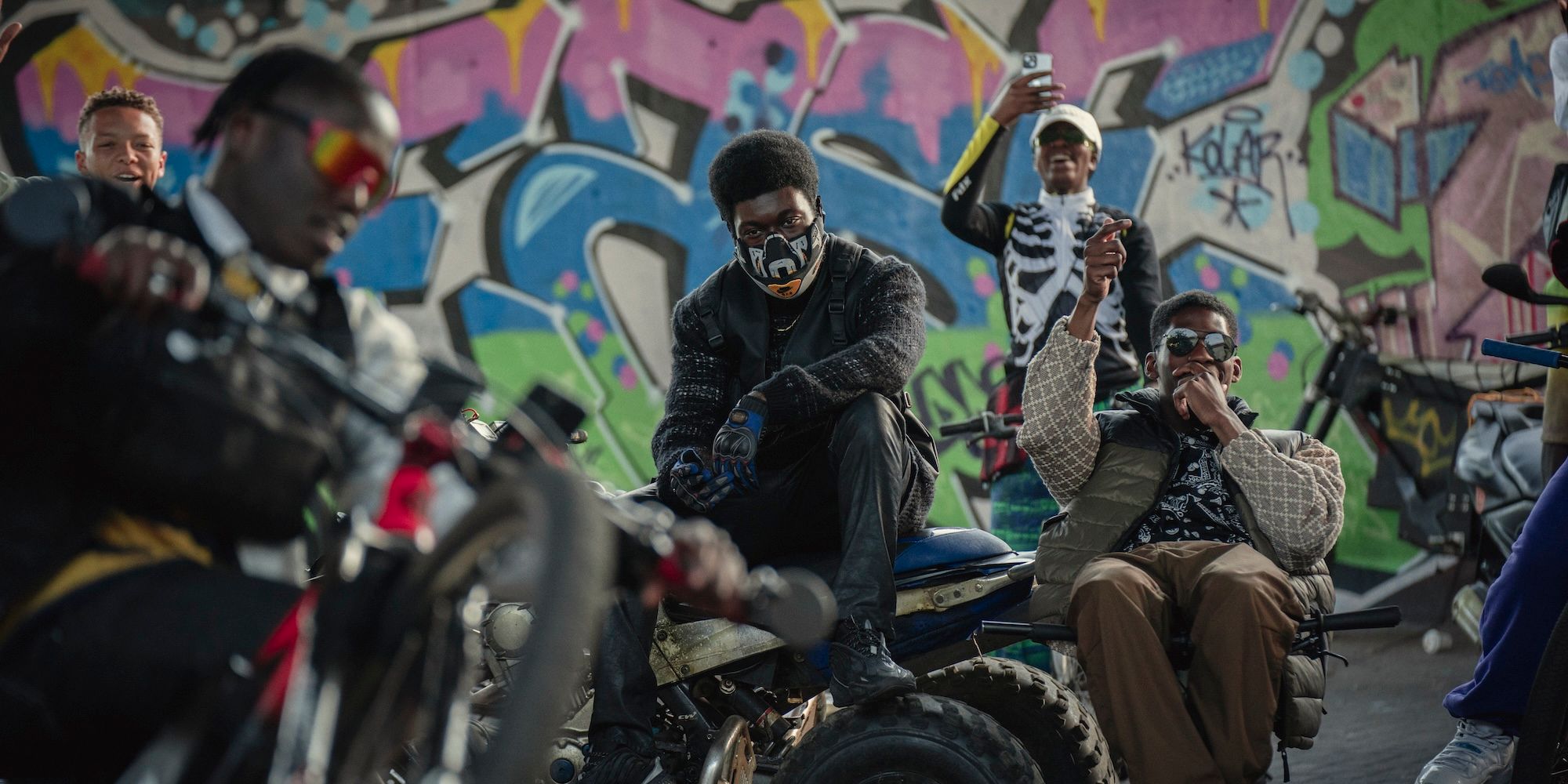 A group of teenage boys ride motorcycles and hang out in front of a graffiti wall in The Kitchen