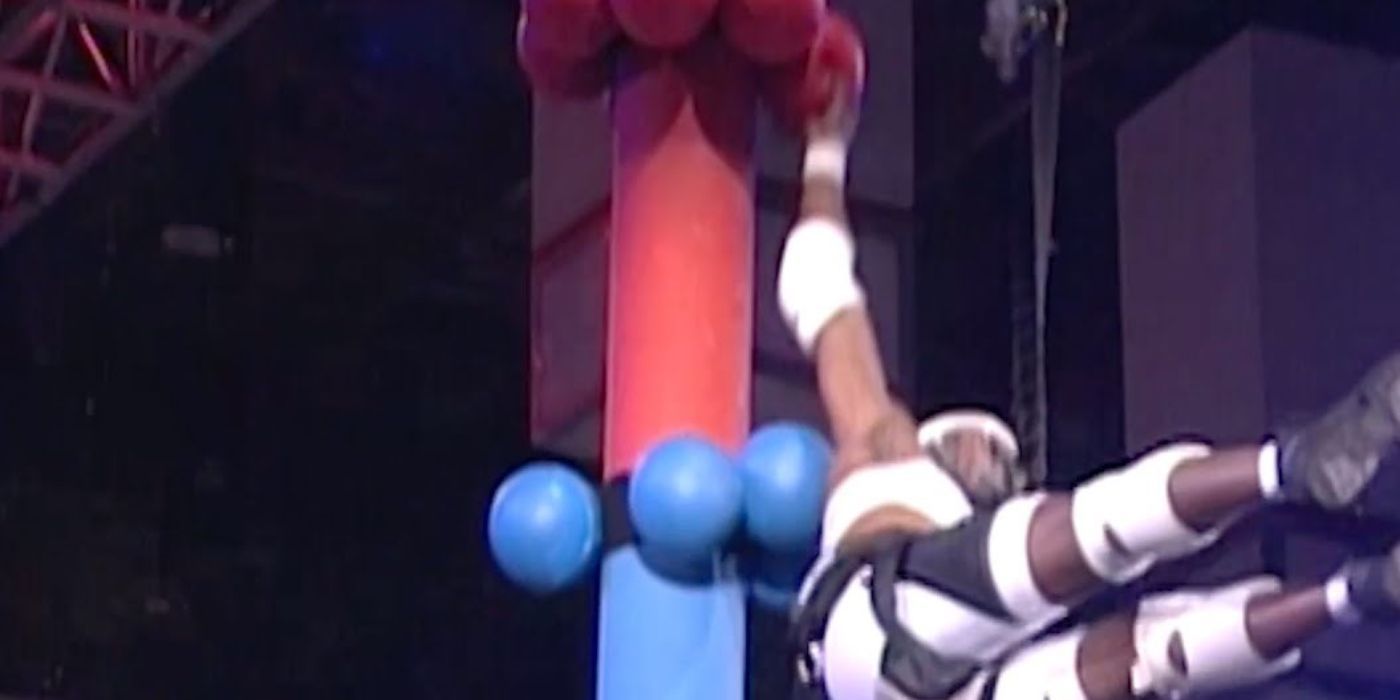 Um homem em uma corda elástica pulando em direção a uma torre com bolas em Swingshot em American Gladiators.