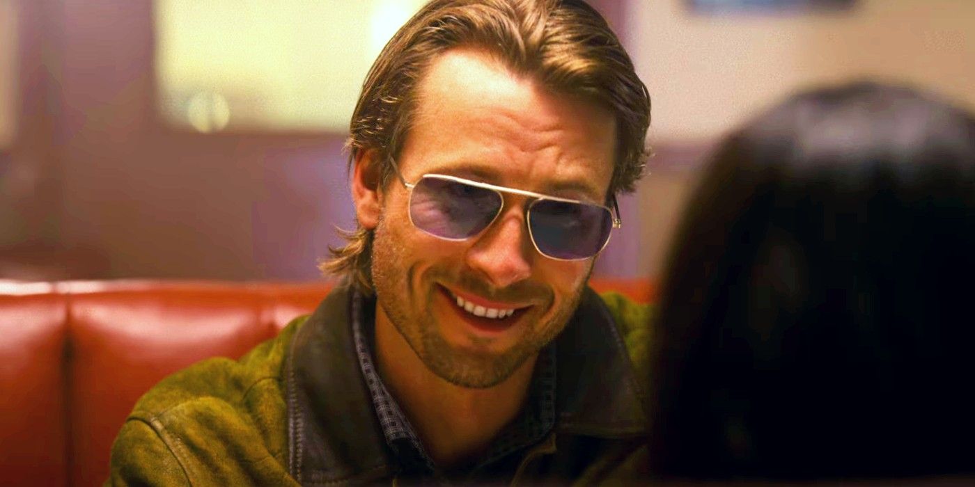 Glen Powell no trailer de Hit Man usando óculos escuros, sorrindo e sentado em uma mesa de jantar