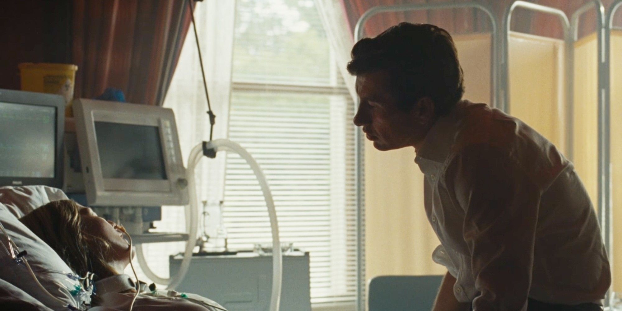 Oliver (Barry Keoghan) leaning over comatose Elspeth (Rosamund Pike) in her hospital bed in Saltburn.