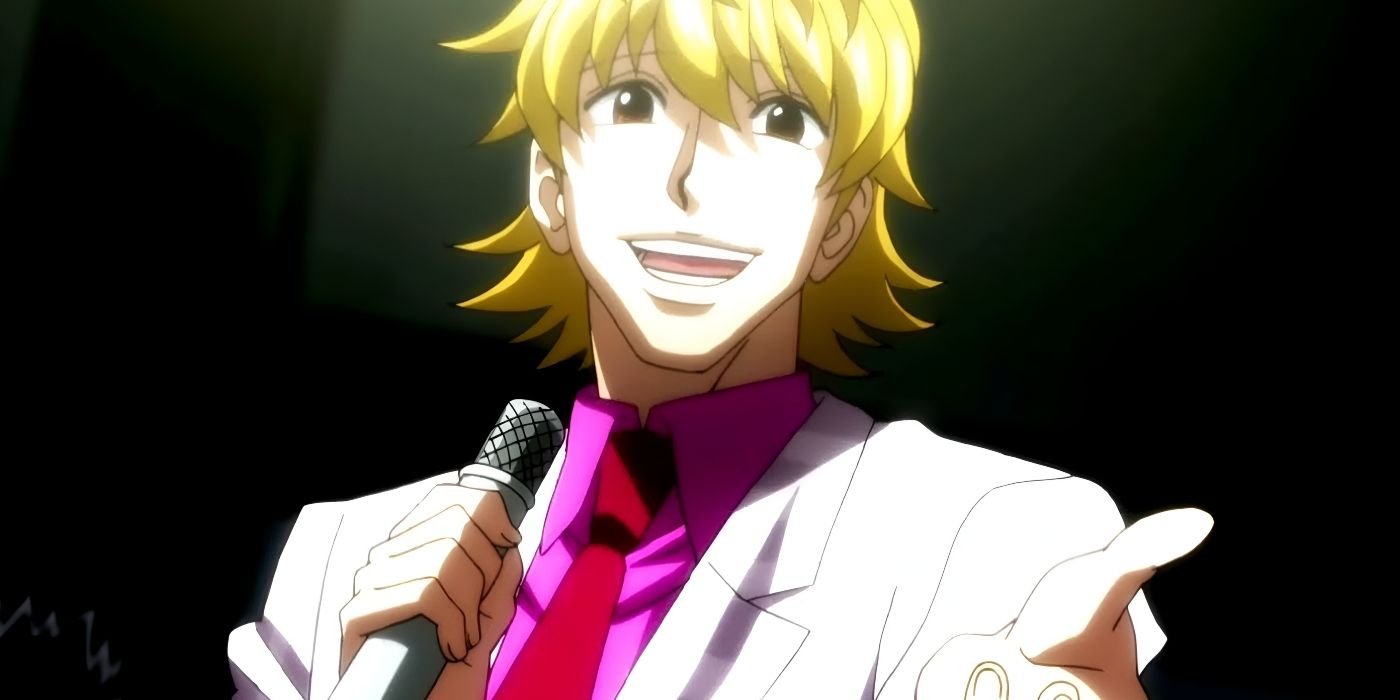 A man with blonde hair smiles as he holds a microphone in one hand and extends his other hand.