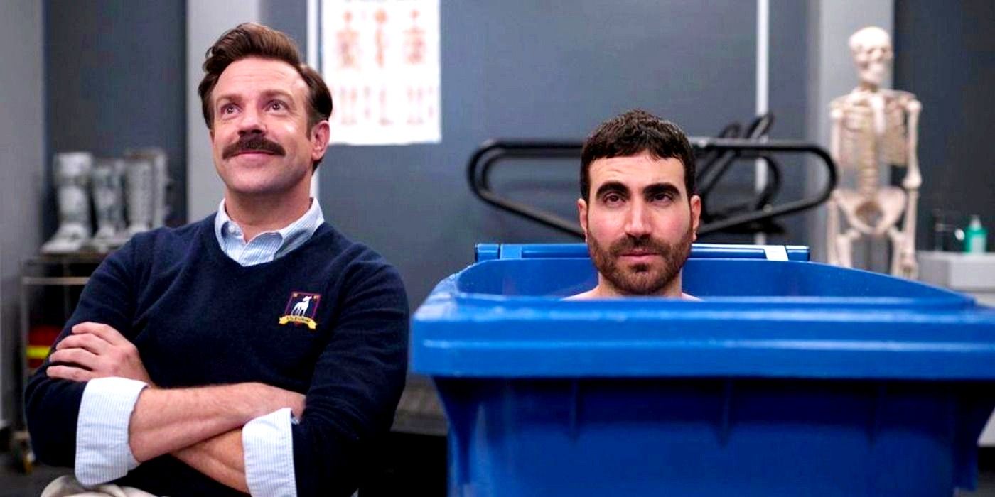 Jason Sudeikis as Ted smiles while Brett Goldstein as Roy is sat in a wheelie bin in Ted Lasso