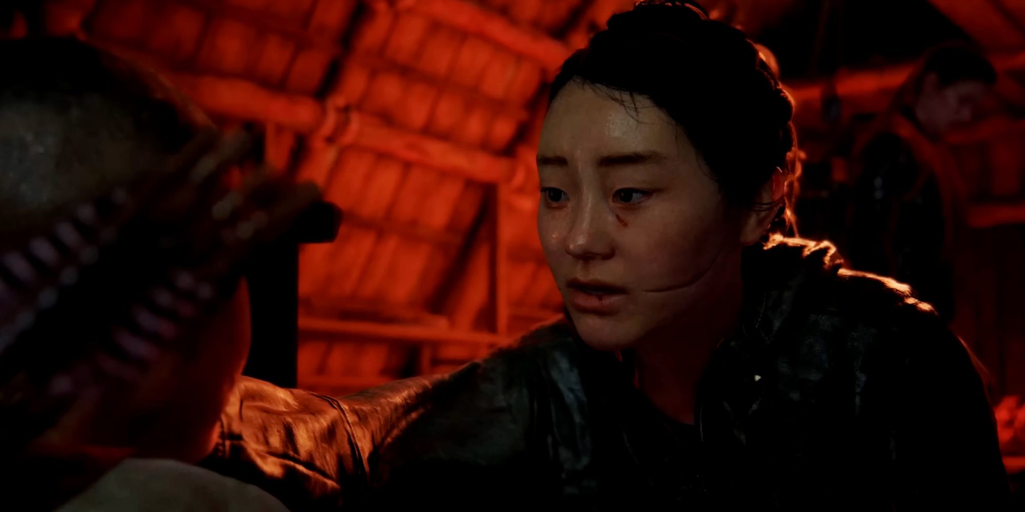 A woman with a deep scar across her cheek is kneeling and consoling a child on the edge of the frame. Another person is standing in the background out of focus in a wooden A-frame building filled with red light.