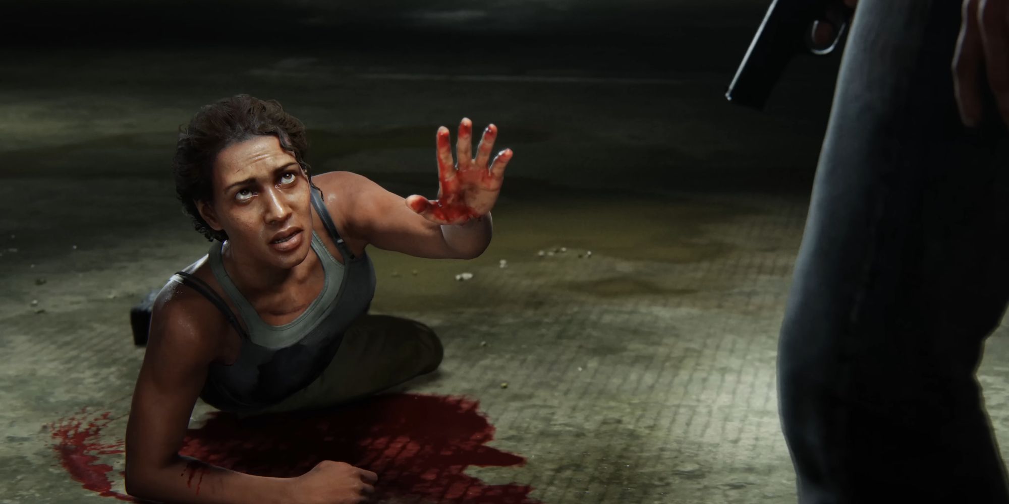 A woman laying on the ground in a pool of her own blood, holding up one hand a looking at someone whose leg can be seen on the edge of the frame. The second person is holding a pistol.