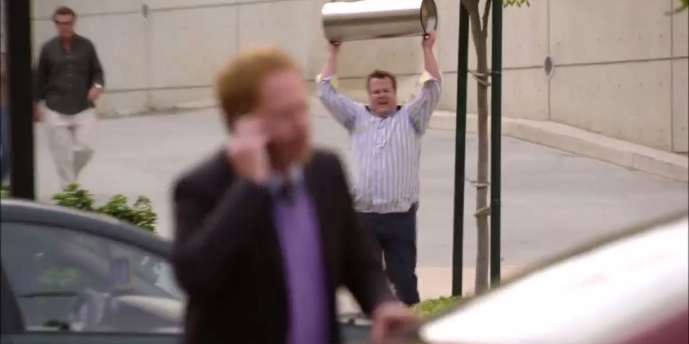 A blurred Mitch on the phone to 911 while Cam runs towards the car screaming with a trash can in the Modern Family episode Run For Your Wife