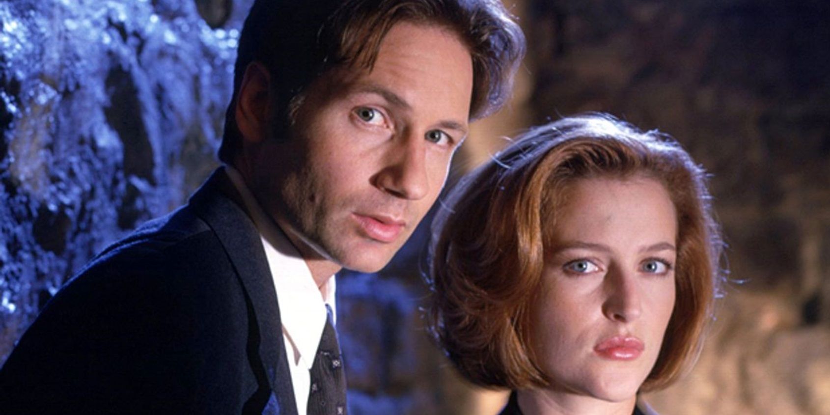 Mulder (David Duchovny) and Scully (Gillian Anderson) stand against a stone wall in a promotional image for The X-Files