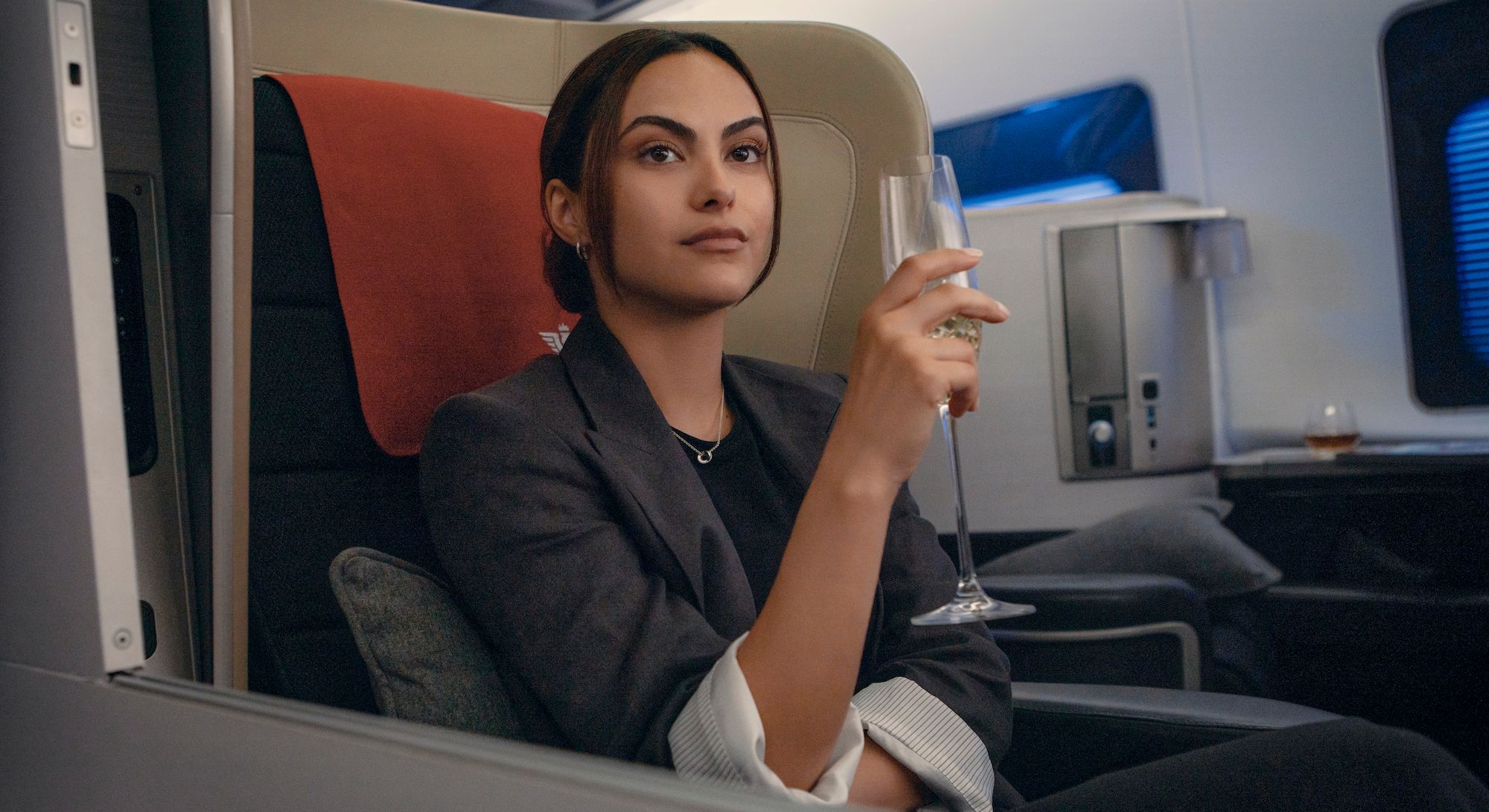 Ana (Camila Mendes) holds a glass of champagne on a plane in Upgraded