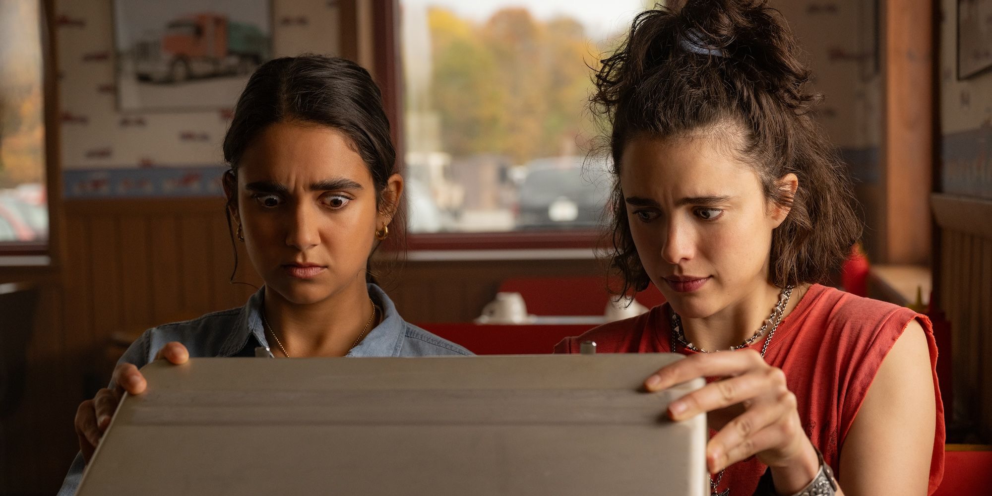Geraldine Viswanathan and Margaret Qualley as Marian and Jamie looking shocked opening a briefcase in Drive-Away Dolls