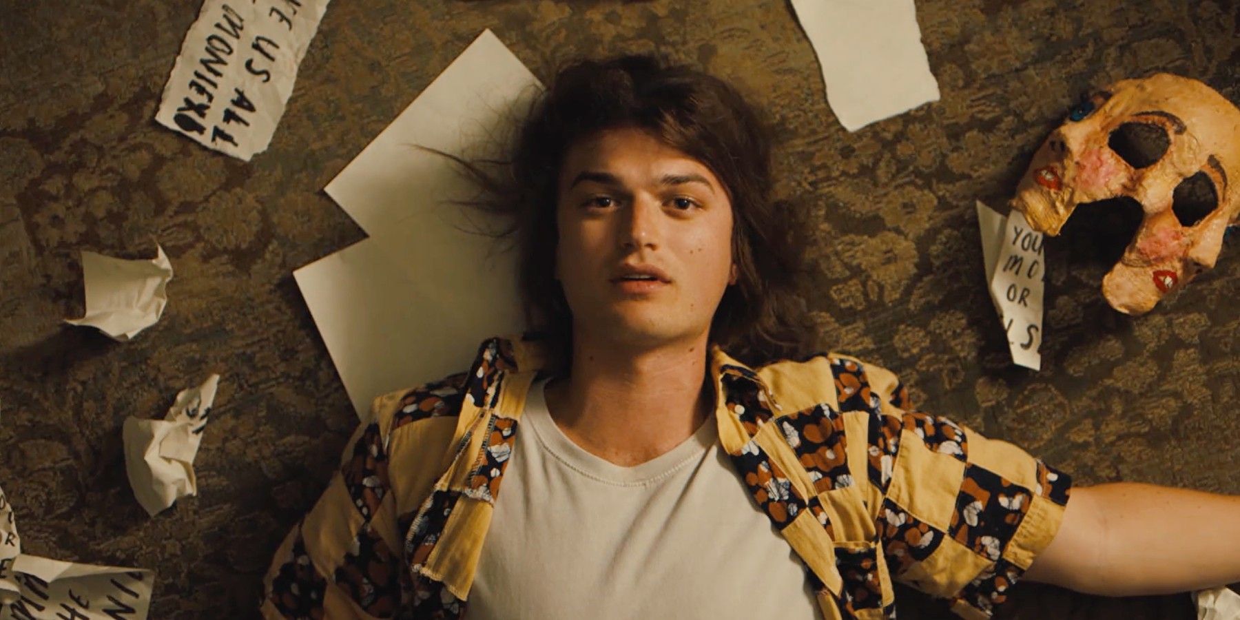 Joe Keery lying on the ground surrounded by scrap papers and a mask in Marmalade