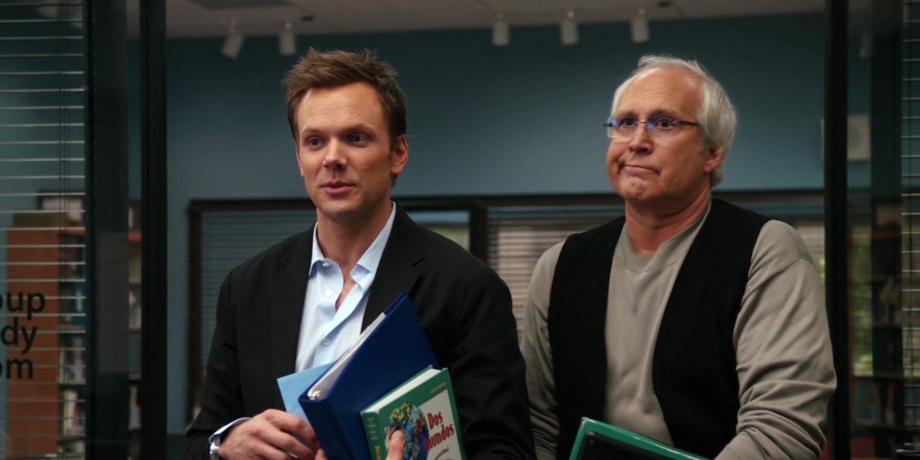 Joel McHale and Chevy Chase standing in the doorway of the study group room holding books in Community