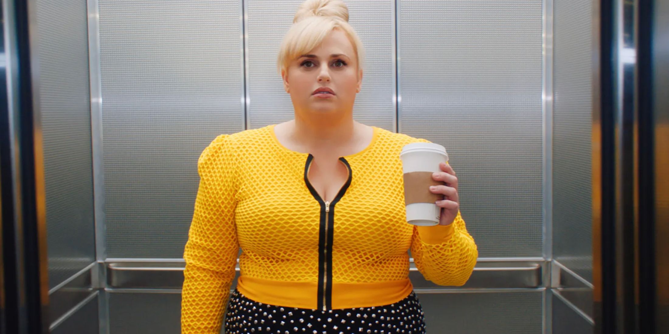 Rebel Wilson com uma blusa amarela brilhante e segurando uma xícara de café no elevador em Isnt It Romantic