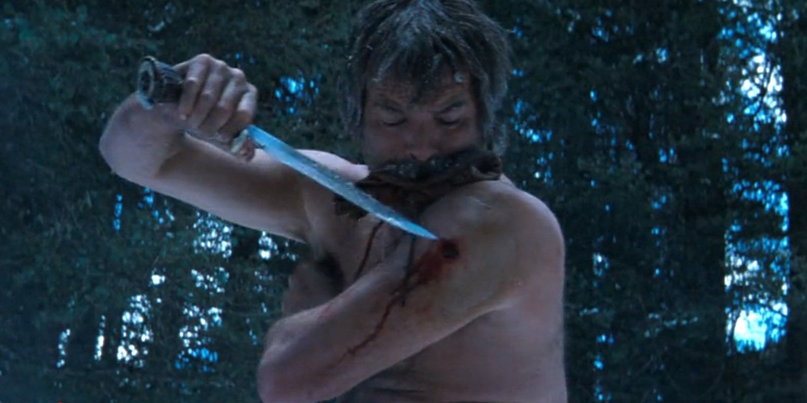 A shirtless Gideon (Pierce Brosnan) digs a bullet out of his arm with a hunting knife while biting on a rag in Seraphim Falls