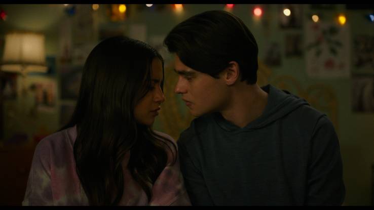 Aza and Davis sit in a dark bedroom with lights behind them. He leans in to kiss her as she closes her eyes.