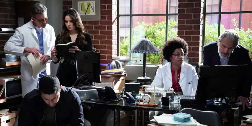 NCIS team members, Palmer, Knight, Torres, Hines, and Parker, working at the office. Parker and Hines are looking at a screen together. Knight and Palmer are looking at notes together. Torres is working on his own.